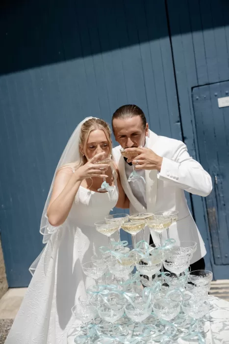 Bride and groom toasting champagne at a coupe glass tower outside Château Canet, with blue wooden doors creating a striking backdrop.