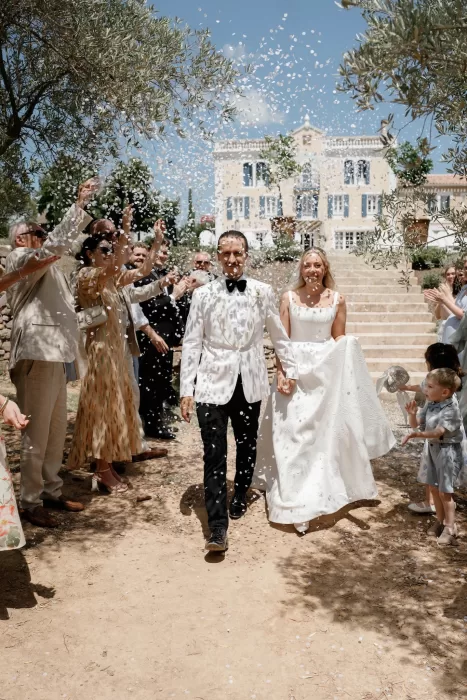 Newlyweds walking through a shower of white confetti at Château Canet, guests applauding beneath olive trees with the château façade rising behind them.