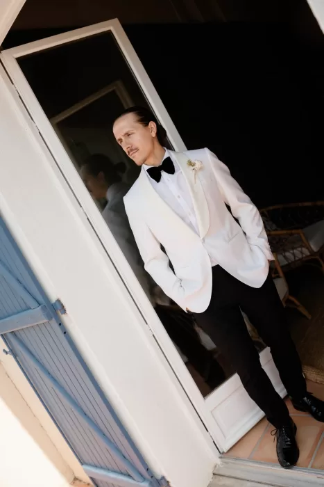 Groom in a white tuxedo jacket and black bow tie standing by blue shutters at Château Canet, captured in warm natural light before the ceremony.