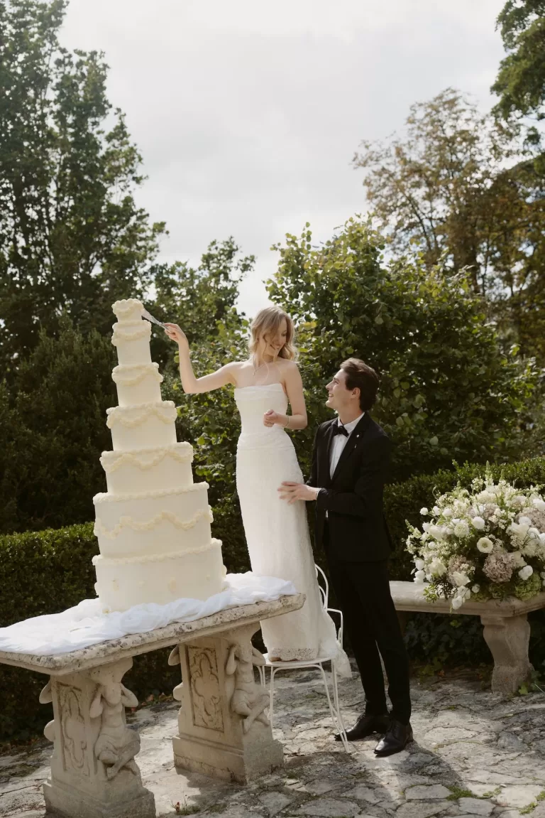 Bride cutting a tall white wedding cake outdoors at Château du Prieuré d’Evecquemont as the groom looks on.