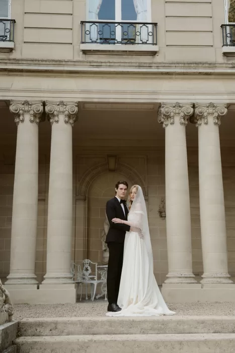 Bride and groom standing beneath the grand columns of Château du Prieuré d’Evecquemont, framed by classical architecture.