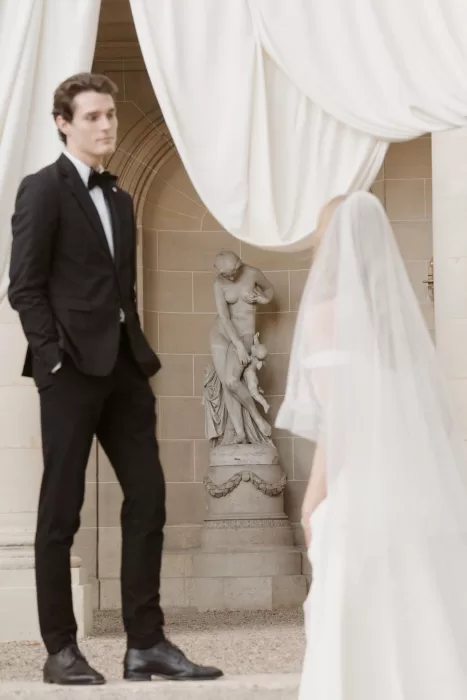 Bride and groom meeting beneath soft drapery near a stone statue at Château du Prieuré d’Evecquemont.