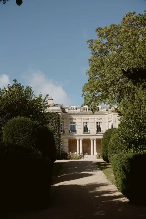Tree-lined gravel path leads towards the elegant façade of Château du Prieuré d’Evecquemont on a clear day.