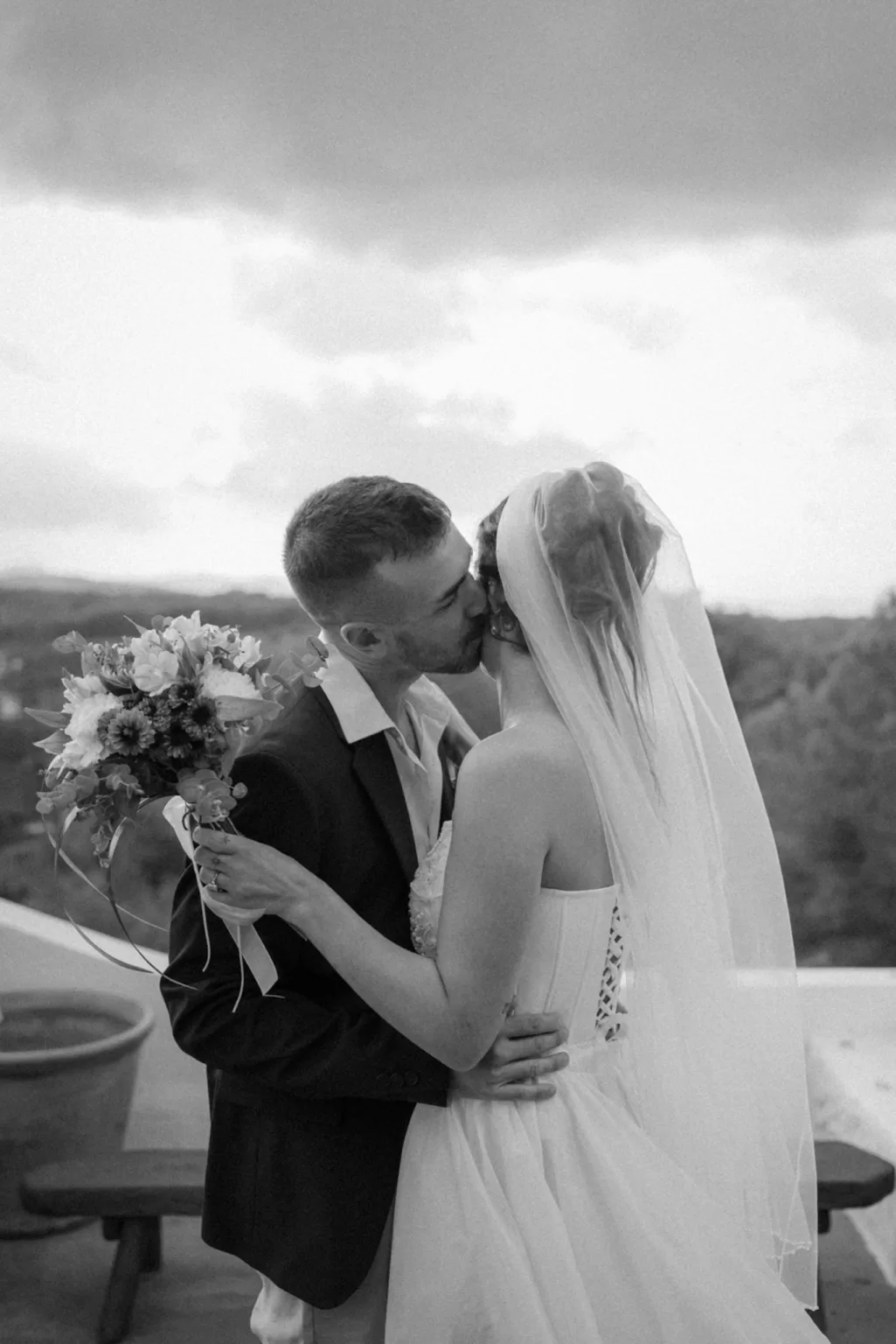 Romantic kiss on a terrace with mountain views, bride holding bouquet as veil flows softly, photographed by Clarissa Sofía Studio.