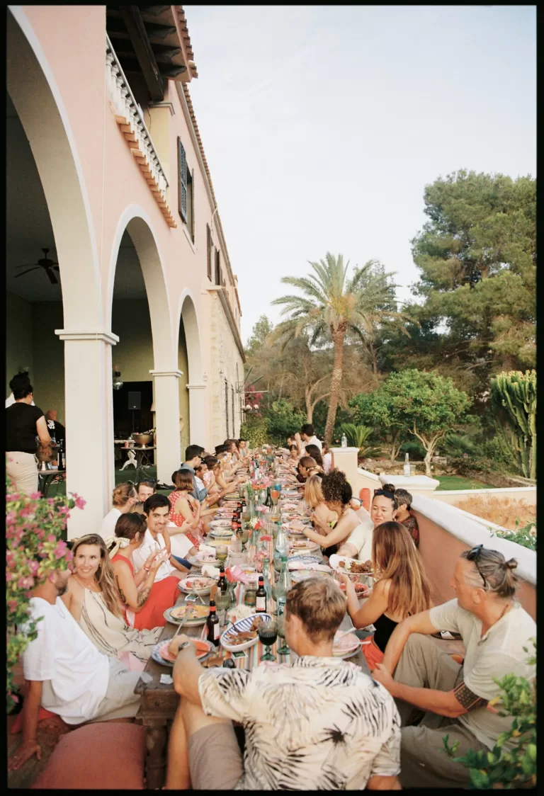 Guests gathered along a long outdoor reception table beside Mediterranean arches, warm sunset dinner captured by Clarissa Sofía Studio.