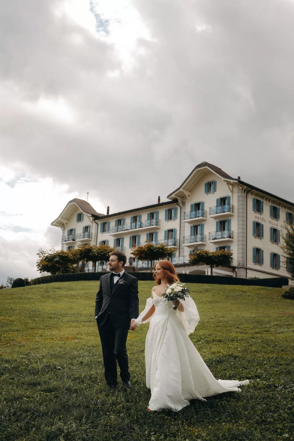 Newlyweds walk hand in hand across the lawn at Hotel Villa Honegg, the bride holding a white bouquet.