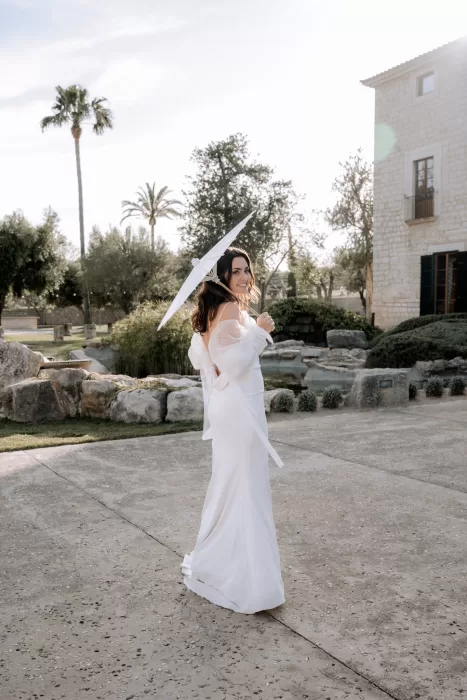 Bride walking through the gardens of Finca Morneta holding a white parasol, sunlight filtering through palm and olive trees.