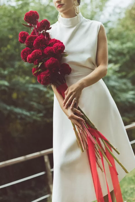 Bride holding deep red bouquet with trailing ribbons on terrace at Hotel Ristorante La Darsena surrounded by greenery.