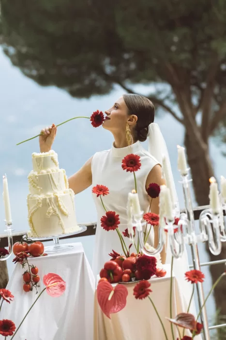 Bride holding a red gerbera beside tiered wedding cake on terrace at Hotel Ristorante La Darsena overlooking Lake Como.