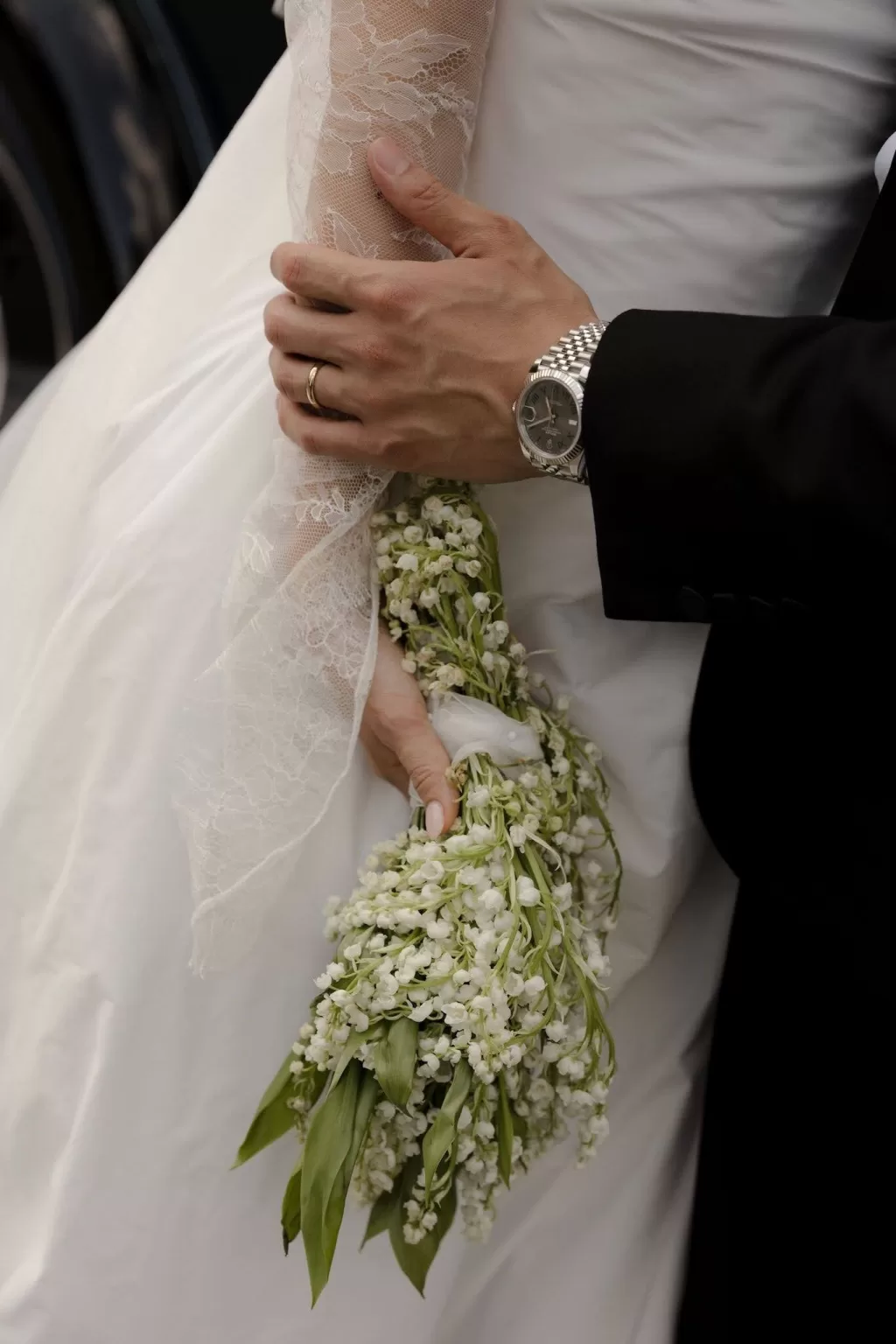 Close-up of the groom’s hand resting on the bride’s lace sleeve, her lily of the valley bouquet held delicately.