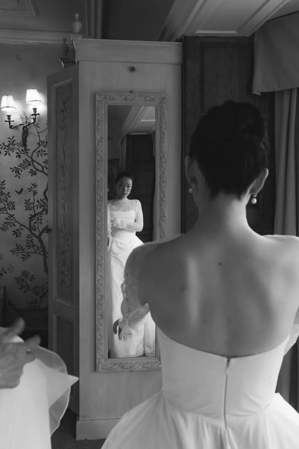 The bride admires her gown in a vintage mirror at Manor House, surrounded by elegant wallpaper and soft light.