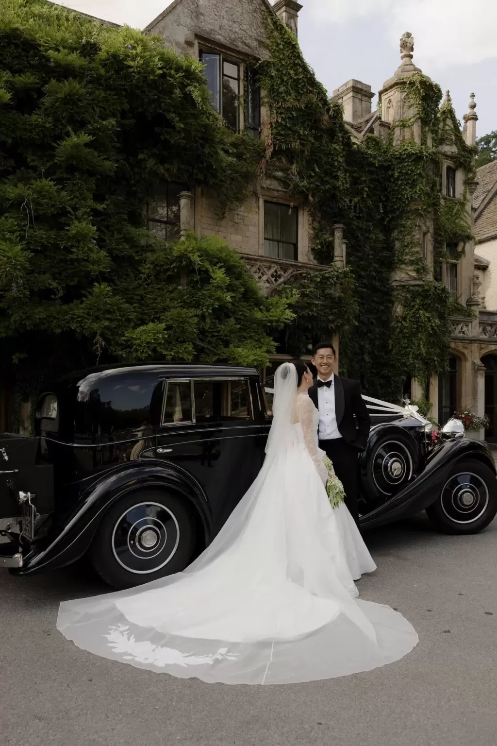 Bride and groom pose beside a vintage black car in front of the ivy-covered Manor House.