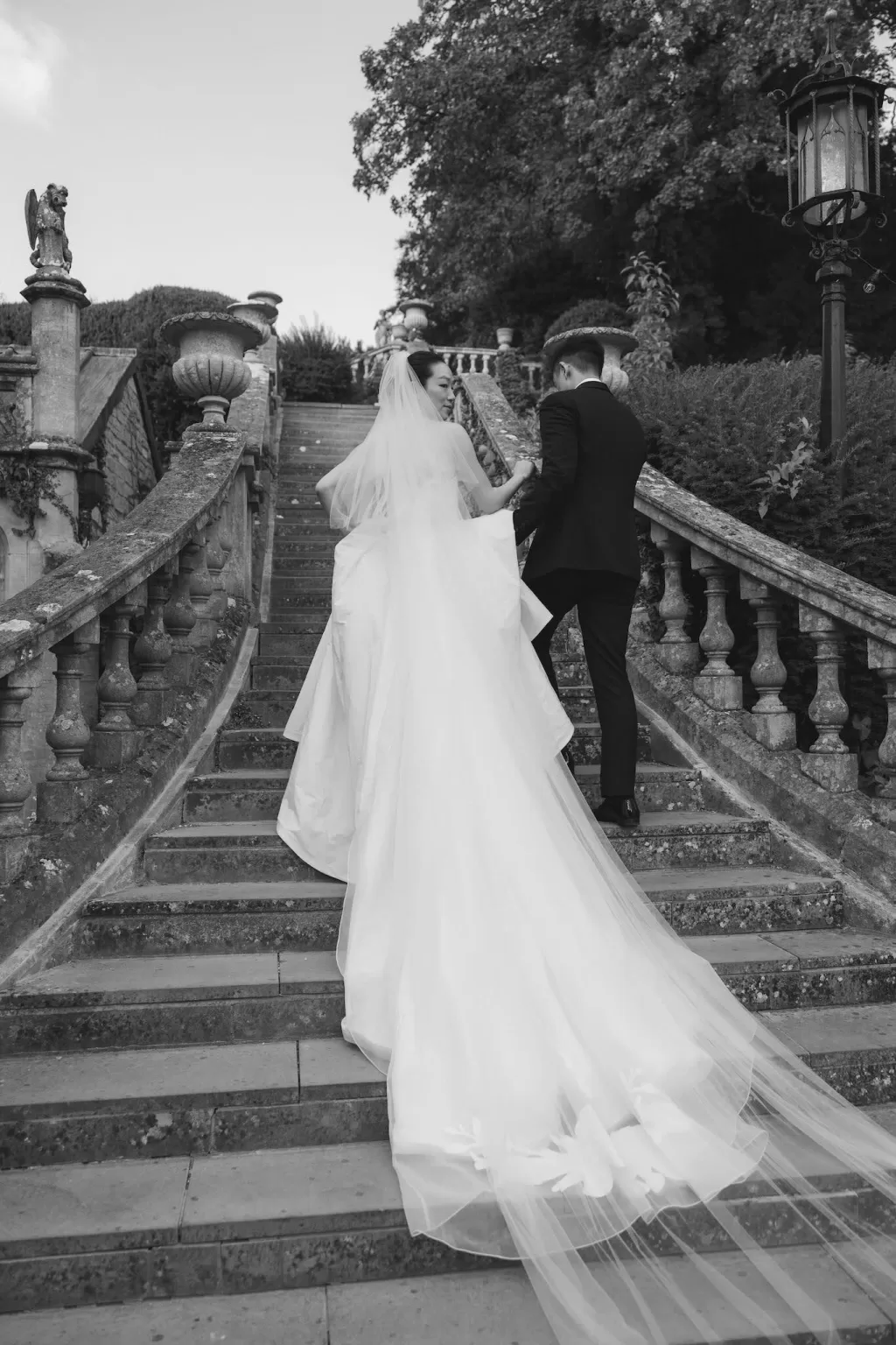 The couple walk hand in hand up the grand stone staircase of Manor House, her veil flowing behind in motion.