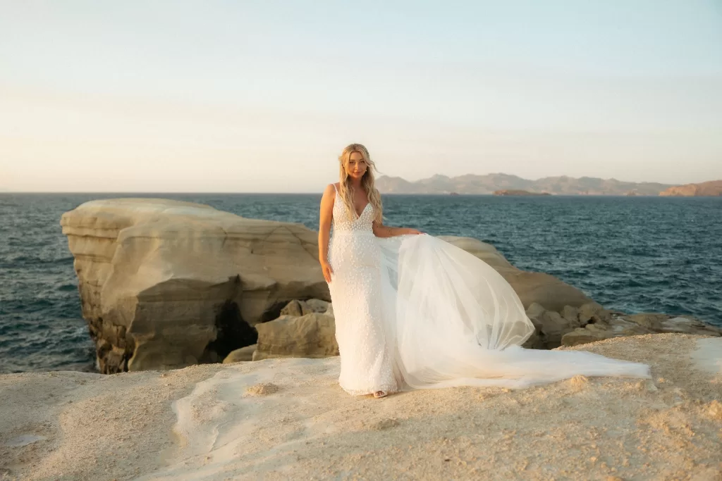 Bride standing on golden coastal rocks near Milos Cove with the Aegean Sea behind her.