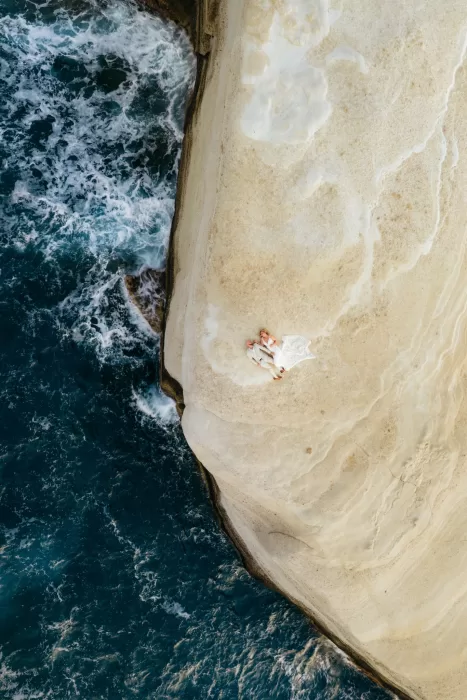 Couple lying on pale rock cliffs above deep blue sea near Milos Cove, captured from above.