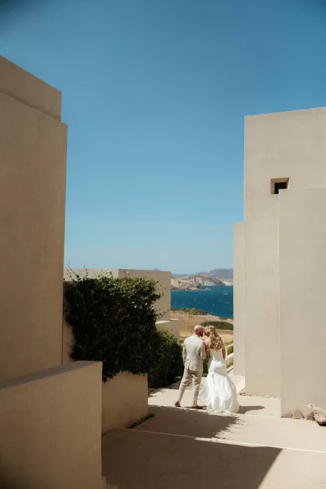 Bride and groom walking between modern cream villas at Milos Cove overlooking the sea.