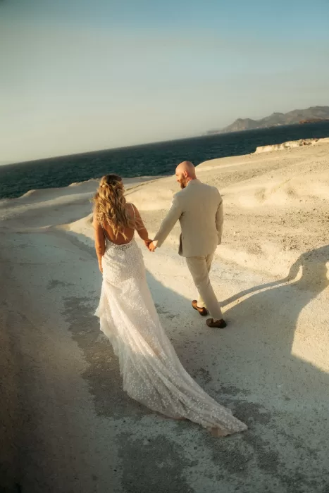 Bride and groom walking hand in hand across golden rock formations near Milos Cove at sunset.