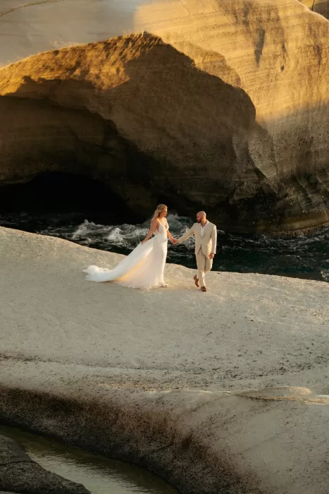 Bride and groom walking hand in hand across golden rock formations near Milos Cove at sunset.