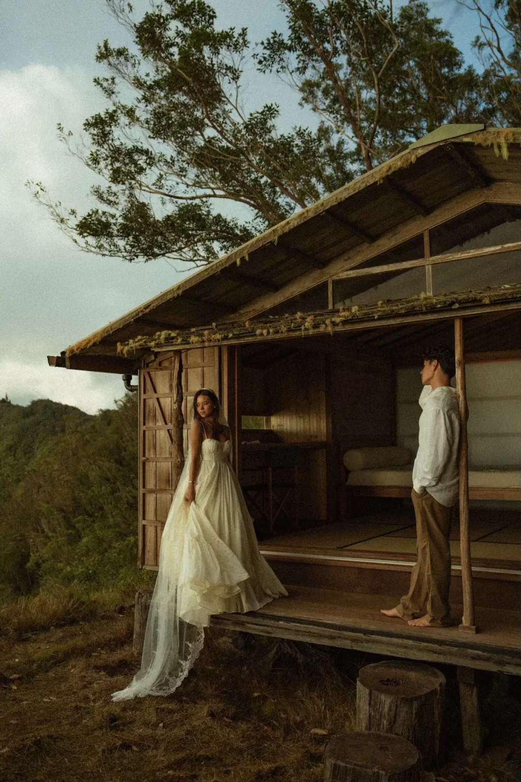 Bride standing on the wooden porch of Ossipoff’s Cabin as the groom looks on, surrounded by forest and soft evening light.