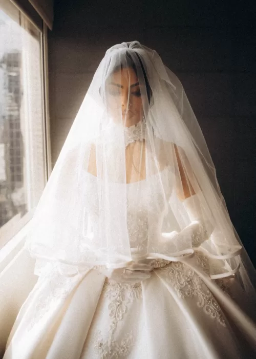 Bride standing by a window at Palacio Metropolitano, her veil softly diffusing natural light over an embroidered satin gown.