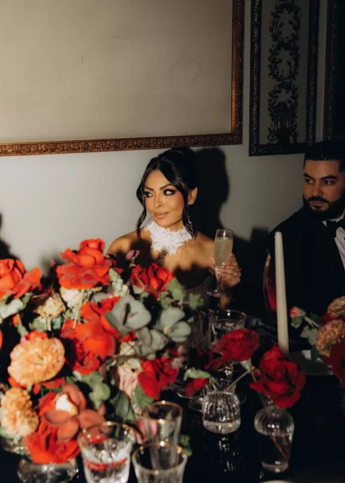 Bride holding a champagne coupe at her candlelit reception table in Palacio Metropolitano surrounded by vibrant red florals.