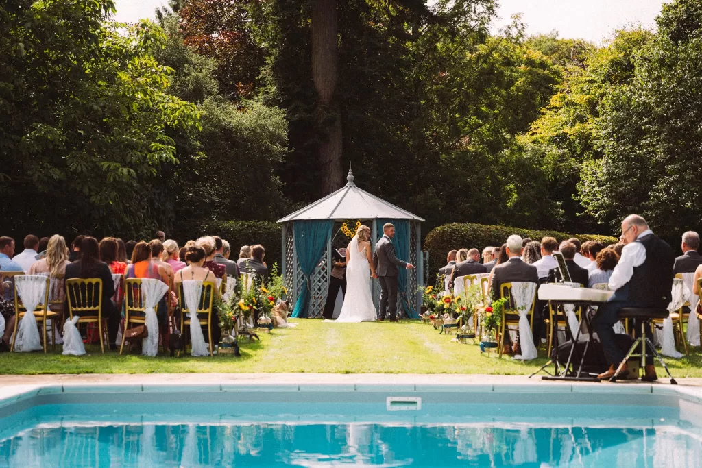 Outdoor wedding ceremony at Scorrier House beside the swimming pool, with guests seated on the lawn facing a garden gazebo.