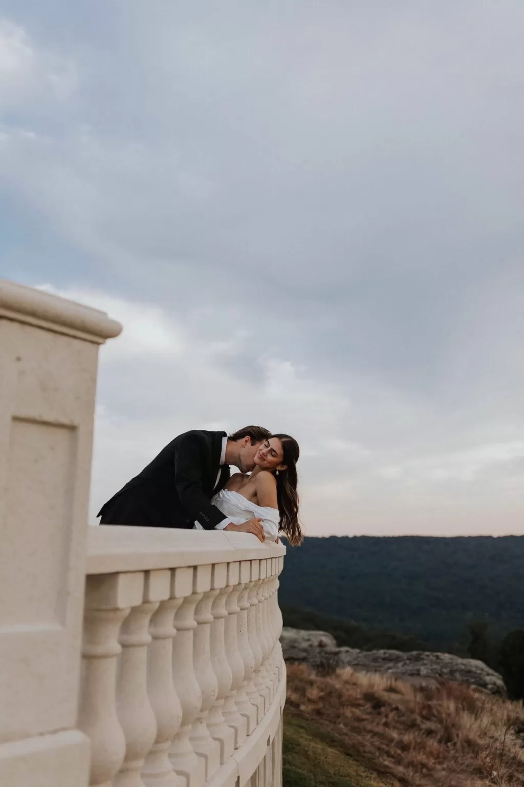 Groom leaning in to kiss his bride on the Stone Haven balcony with dramatic landscape views.