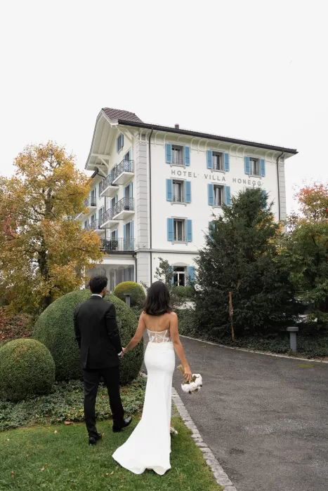 Bride and groom walking toward the iconic white façade of Villa Honegg surrounded by autumn trees.