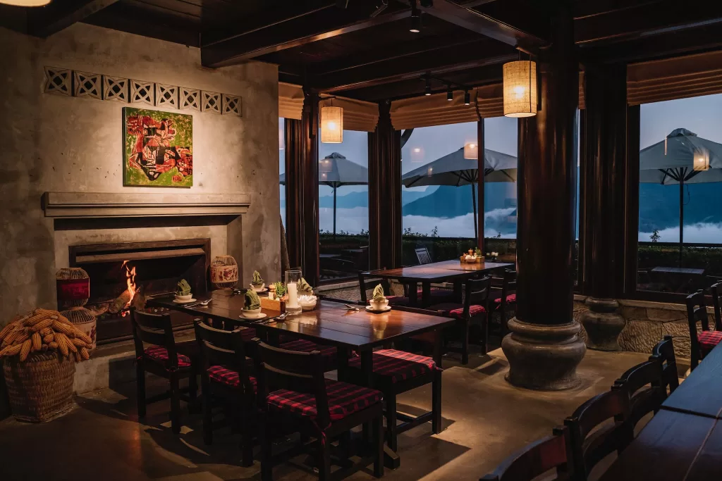 Warm restaurant interior at Ville De Mont Mountain Resort with wooden tables, fireplace, and panoramic windows overlooking clouds.