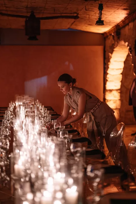 Event stylist arranging glassware along a long candlelit reception table inside Wadi Rum stone venue.