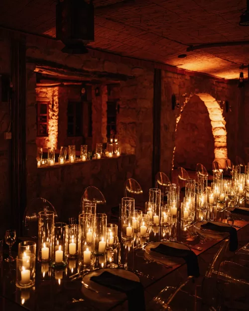 Romantic candlelit wedding dinner inside stone reception space at Wadi Rum, with rows of glass cylinders and soft amber lighting reflecting on the tables.