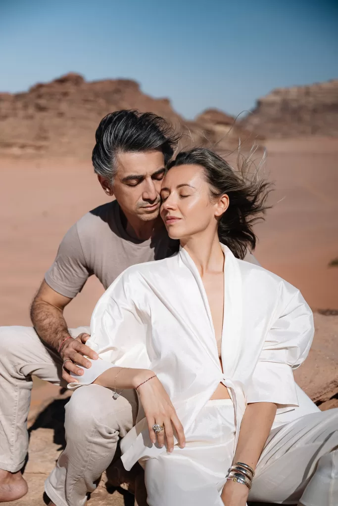 Couple seated closely on sunlit rocks in Wadi Rum desert, wind moving through her white outfit against the sandstone backdrop.