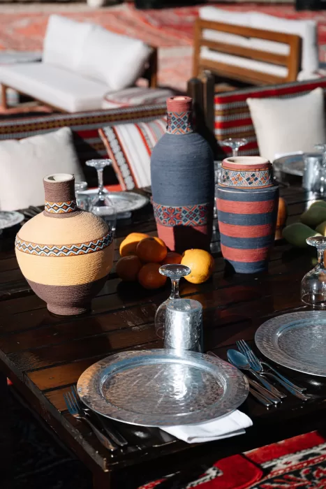 Long wooden reception table set on patterned rugs in Wadi Rum desert, styled with woven vases, silver plates and fresh citrus under warm sunlight.