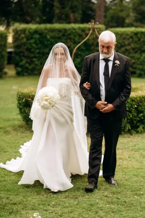 Bride walking with father through garden at Castello di San Fabiano, holding white bouquet under soft veil