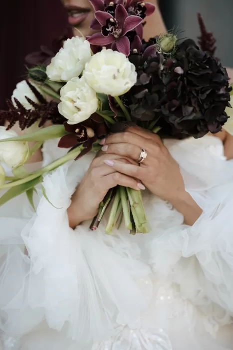 Bride holding a dramatic bouquet of orchids, tulips, and dark florals with her engagement ring visible at Common Goods.