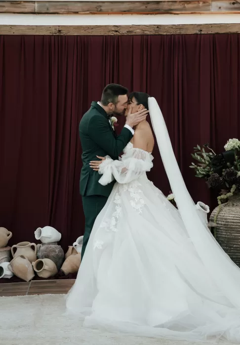 Bride and groom sharing a kiss in front of burgundy drapery and rustic ceramic décor during their wedding at Common Goods.
