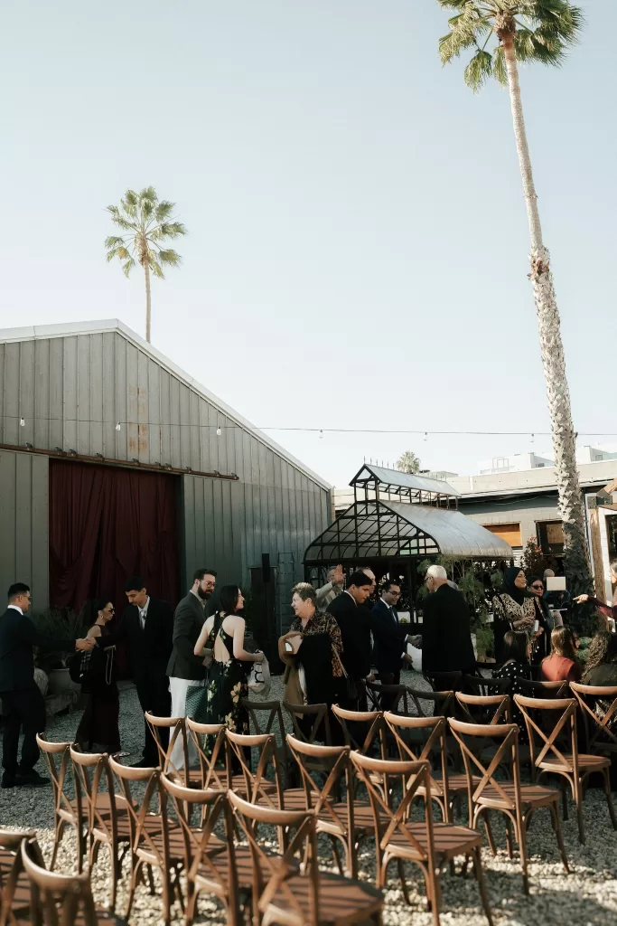 Wedding guests gathering among wooden ceremony chairs outside Common Goods with palm trees and greenhouse in the background.