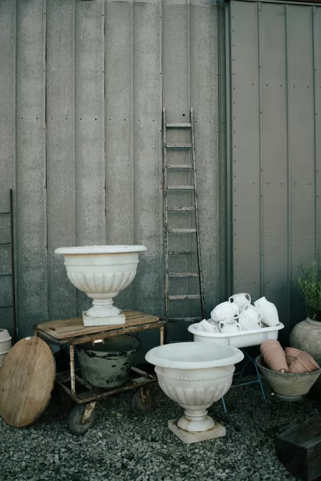 Rustic outdoor display at Common Goods with vintage ladder, ceramic planters, and stacked pitchers against a metal wall.