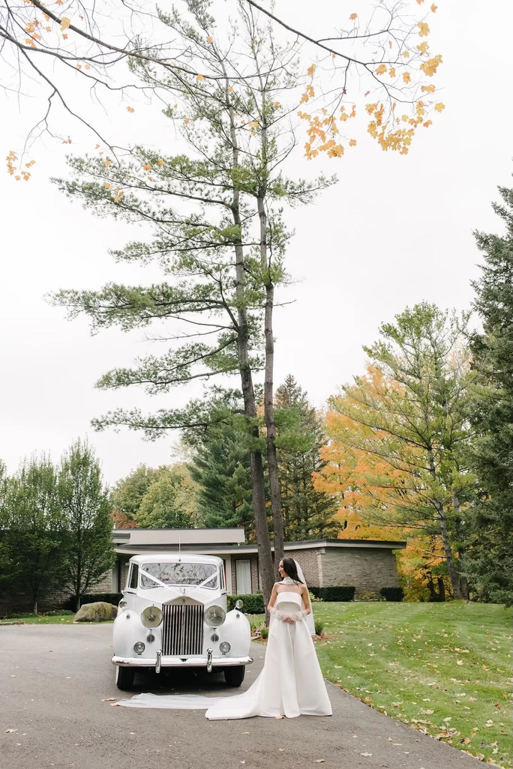 Bride standing beside a classic white car surrounded by autumn trees outside Graydon Hall.