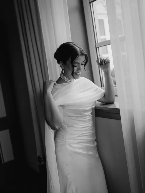 Bride smiling softly by a window in her elegant cape wedding dress at Infante Sagres, lit by natural light.
