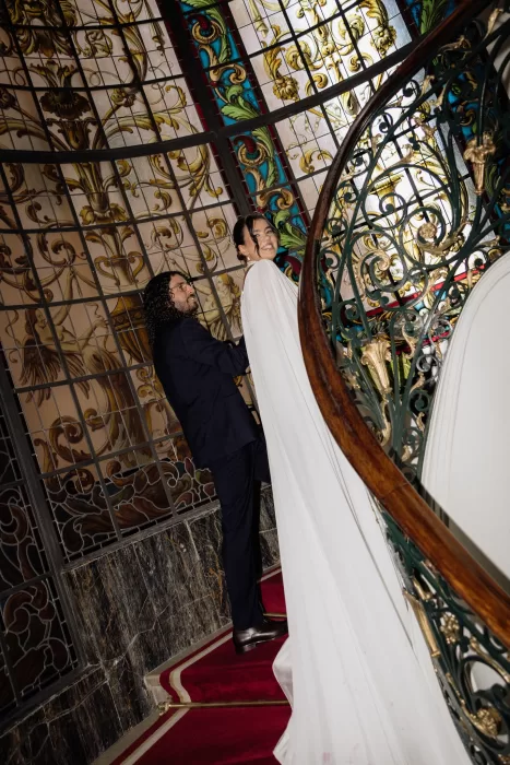 Bride and groom walking up the grand staircase beneath stained glass ceiling inside Infante Sagres.