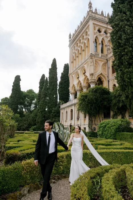Bride and groom walking through manicured gardens at Isola del Garda with historic villa behind