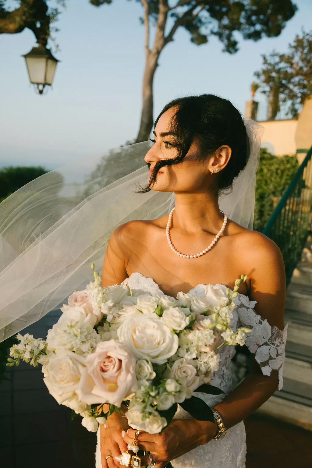 Bride holding white rose bouquet with veil flowing in golden sunset light in Capri