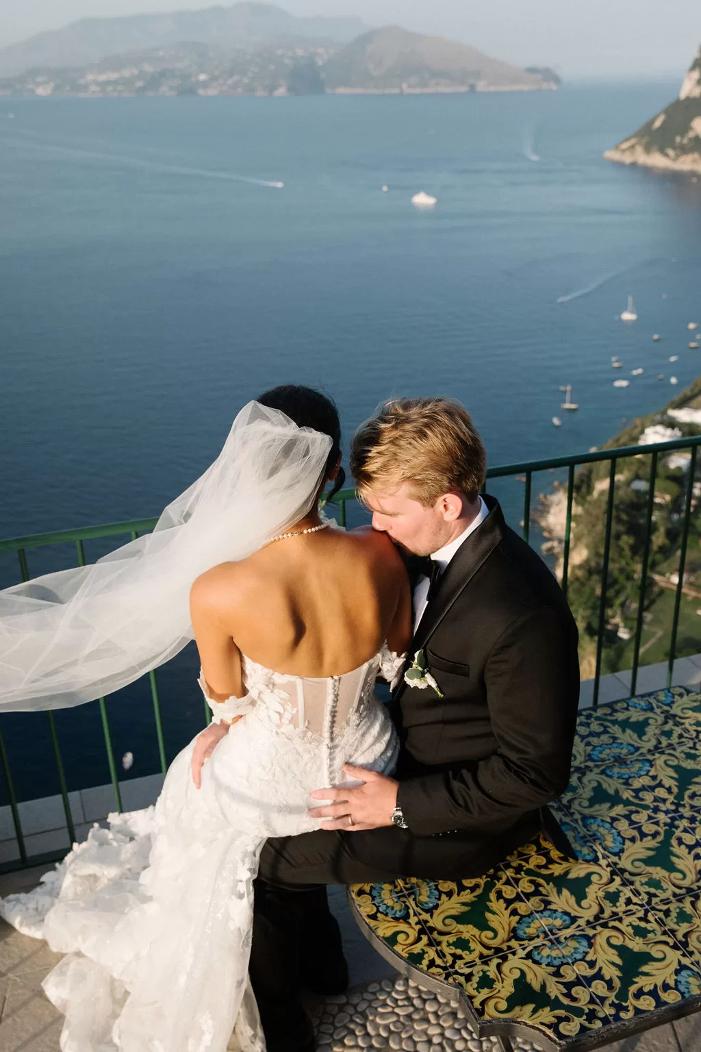 Bride and groom embracing on a balcony overlooking Capri coastline and blue sea