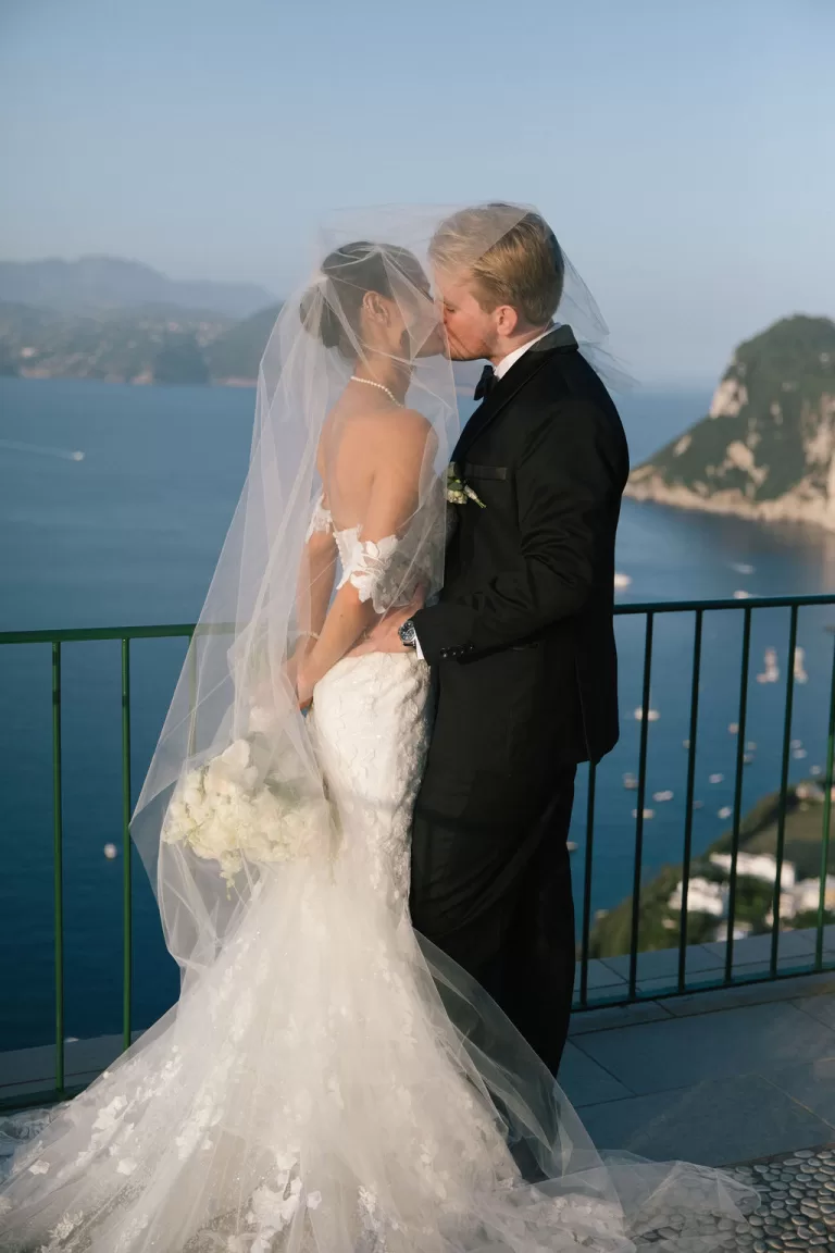 Bride and groom kissing on balcony overlooking Capri cliffs and blue sea