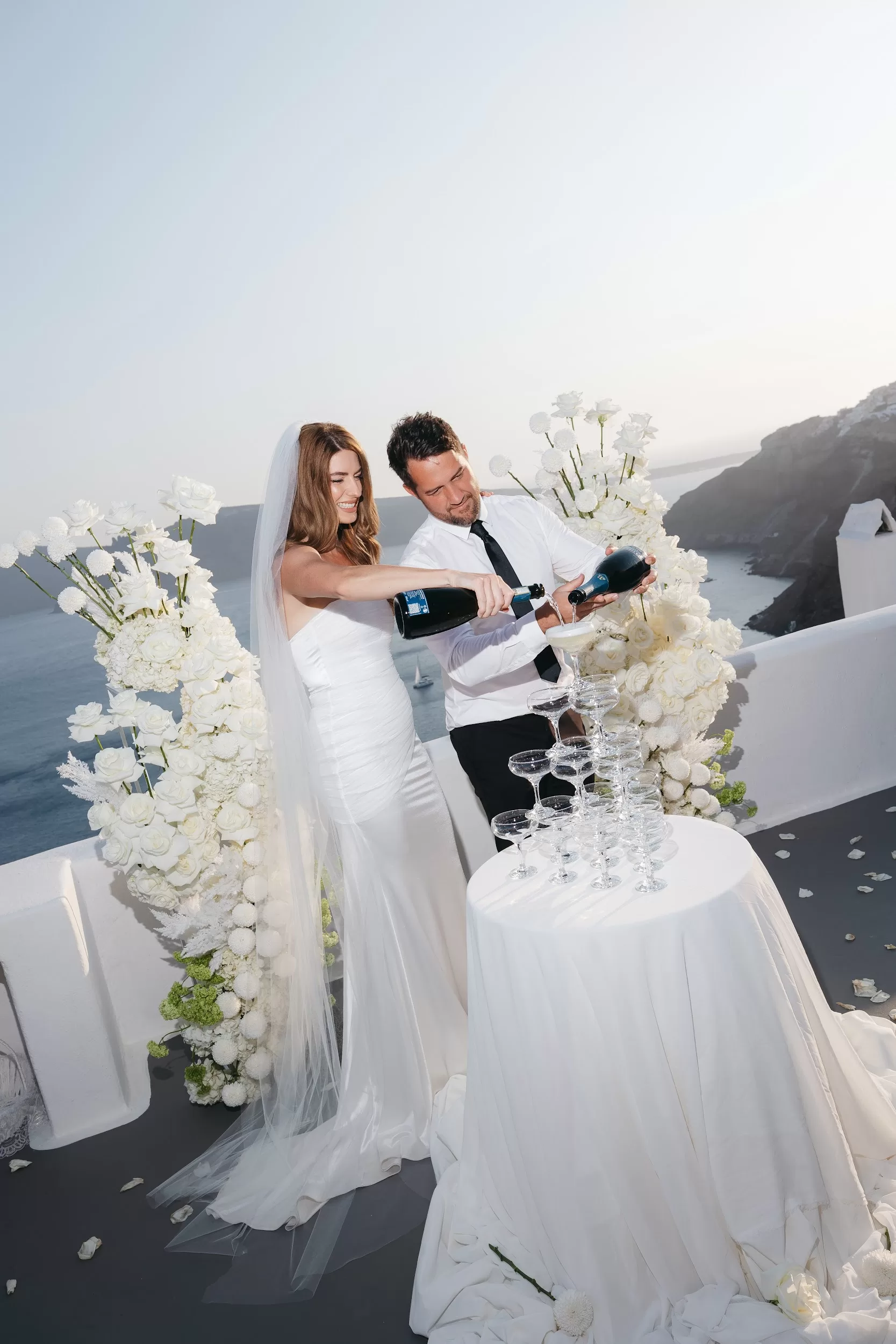 Bride and groom pouring champagne into a coupe glass tower during a Santorini wedding celebration.
