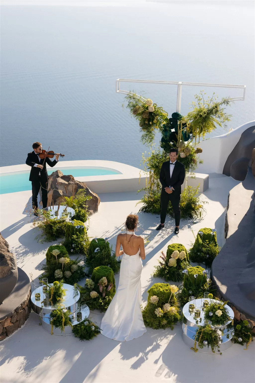 Bride walking toward groom during an outdoor Santorini ceremony surrounded by lush green floral installations.