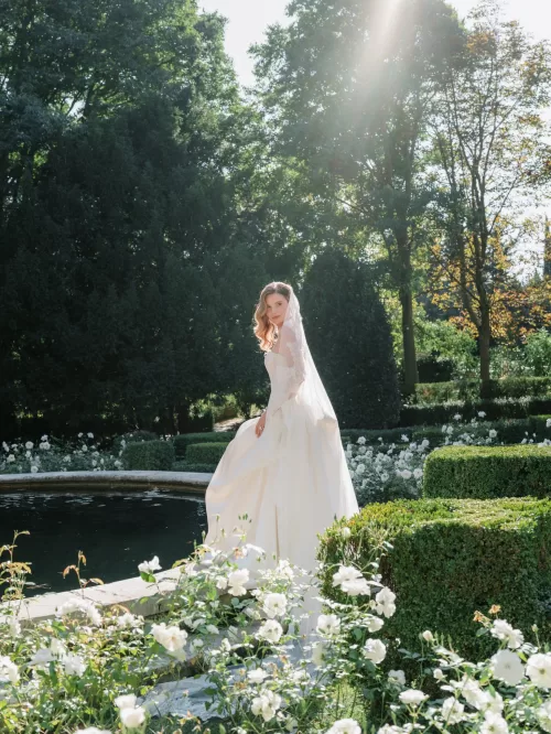 Bride in flowing gown standing beside a garden pond surrounded by white roses at Mas de Chabran.