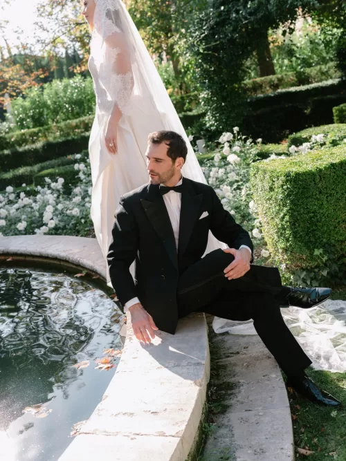Groom seated beside a garden pond while the bride stands in a flowing veil among roses at Mas de Chabran.