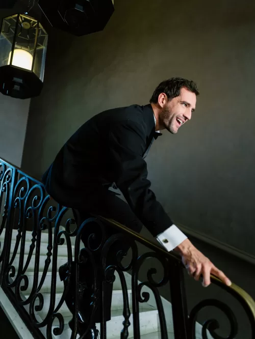 Groom in black tuxedo leaning over an ornate staircase railing inside Mas de Chabran.